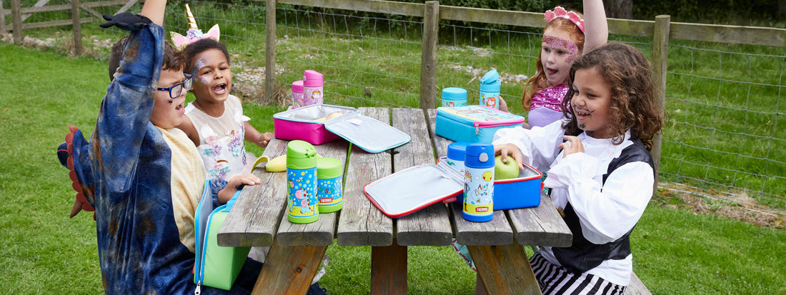 Four children in fancy dress having fun whilst eating a picnic on a park bench and eating and drinking from Thermos flasks