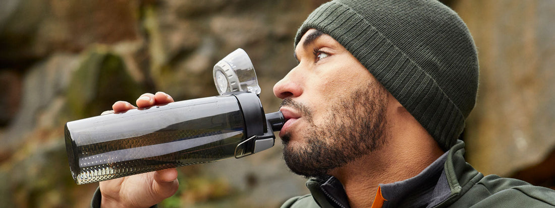 A man with a wool hat drinking from a Thermos Water Bottle with a rocky face background