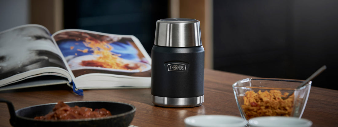 A Thermos Food Flask on a coffee table with a cook book open and the ingredients in dishes