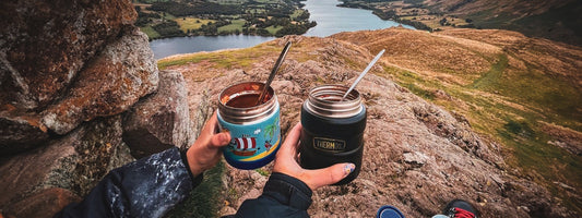 Two peoples hands holding Thermos Food Flasks with food in them with a background view of the countryside and a river