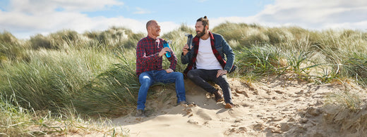 Two men sat in the sand dunes by the sea chatting and drinking from Thermos Water Bottles