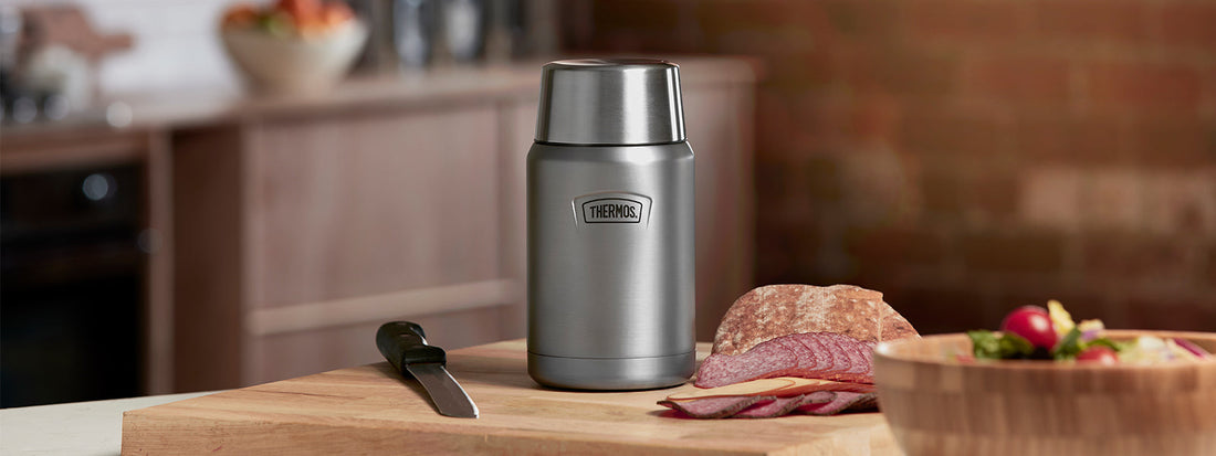 A chopping board in a kitchen setting with some salad and meat being prepared next to a Thermos Food Flask